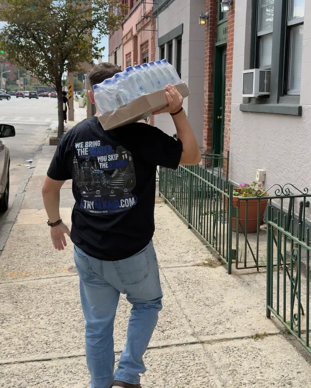 An Alkaid driver carrying a case to a door, his shirt reading: we bring the water, you skip the stairs
